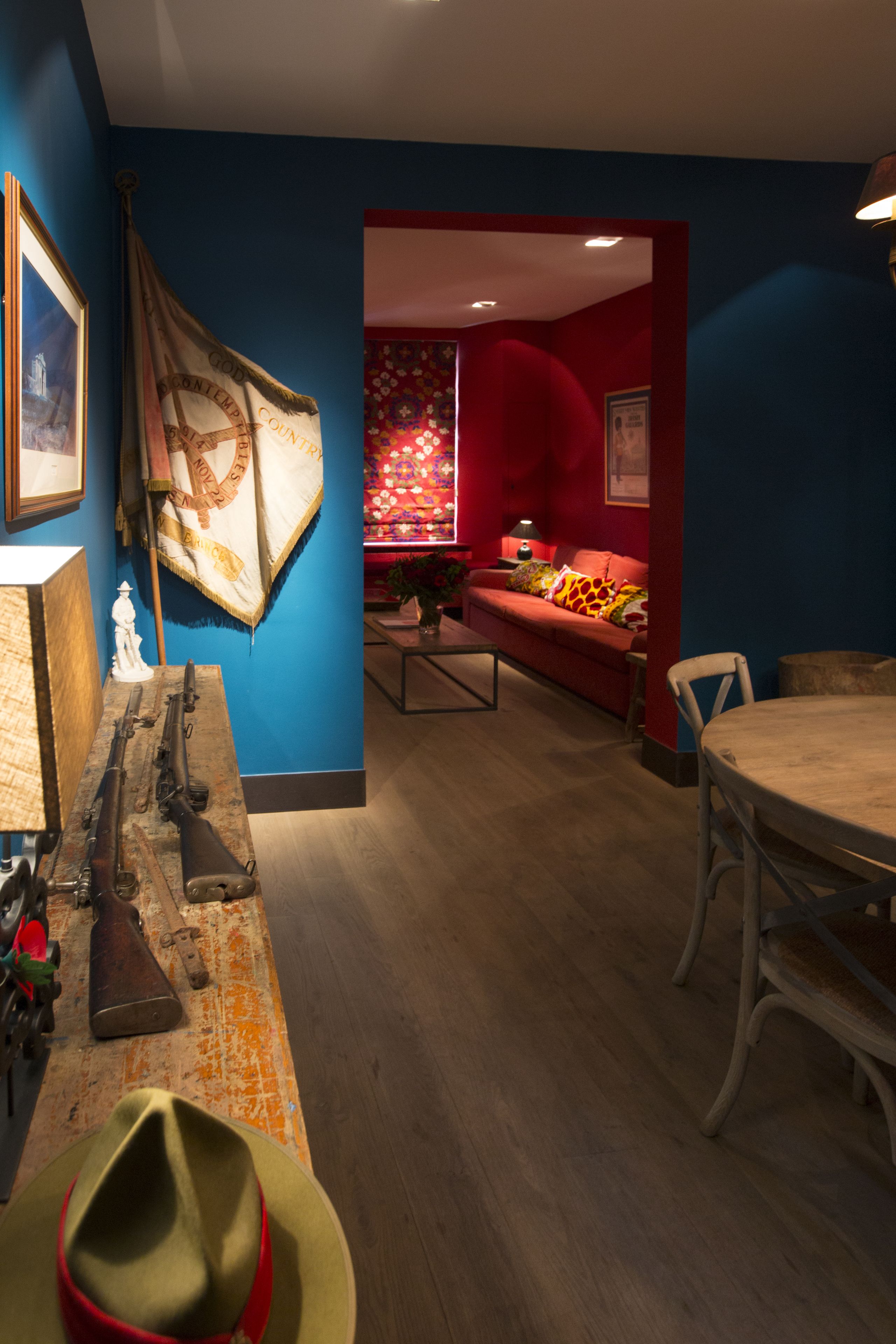 Une pièce avec une table en bois, un canapé rouge et une table à manger. On peut voir des fusils et un drapeau accrochés au mur.