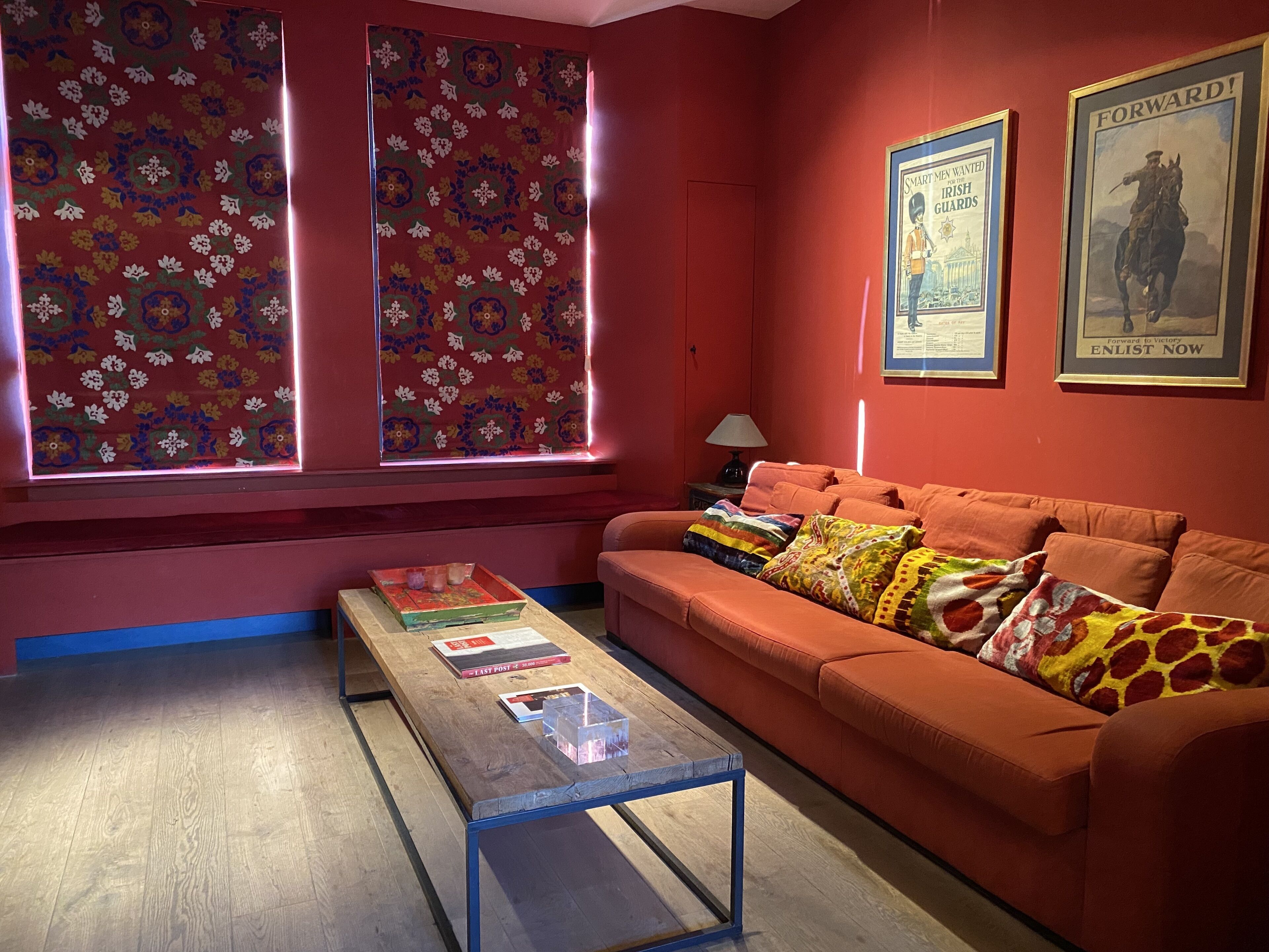 A room with a red wall, a sofa with colorful cushions, a wooden coffee table with books, and two posters on the wall.