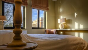 A hotel room with a bed, a wooden table, a lamp, and a window with a view of buildings. - Menin Gate House  1 (Ypres)
