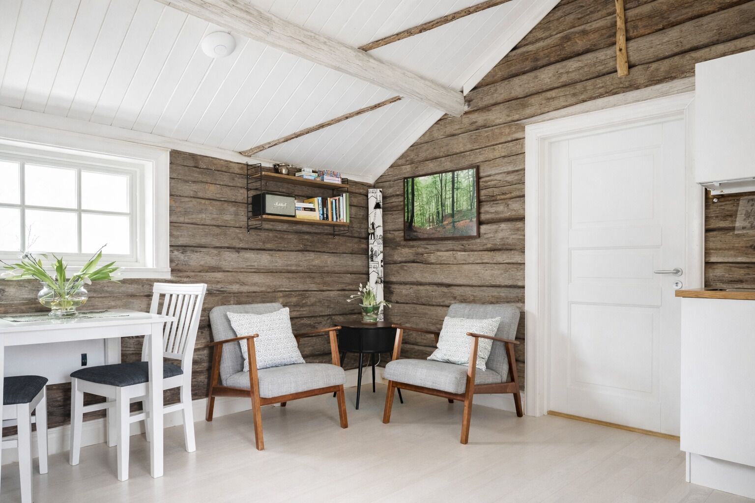 Une pièce chaleureuse avec un mur en bois, une porte blanche, une table à manger avec des chaises et une petite table d’appoint sur laquelle est posé un vase.