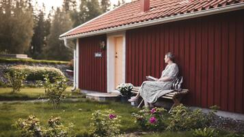 A person sitting on a bench outside a red building with a tiled roof, surrounded by a garden with various plants and flowers.
