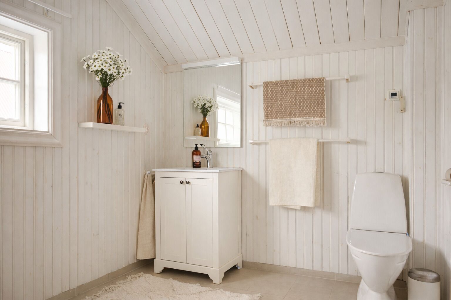 Ein Badezimmer mit weißer Holzvertäfelung, einem weißen Waschtisch mit Spiegel, einer Toilette und einem kleinen Ablagebereich für Toilettenartikel.