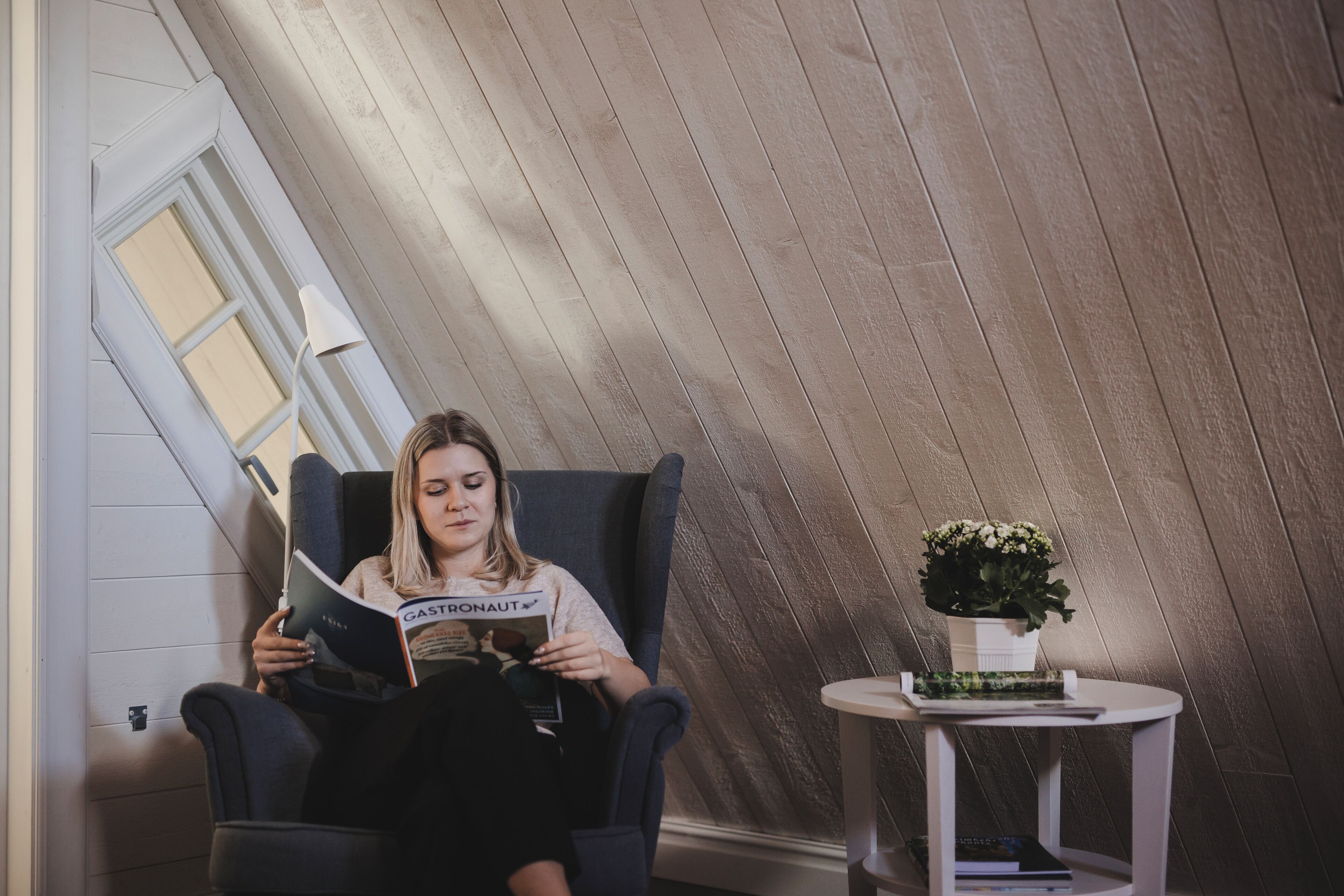 A person sitting in a chair reading a magazine in a room with wooden paneling and a small table with a plant.