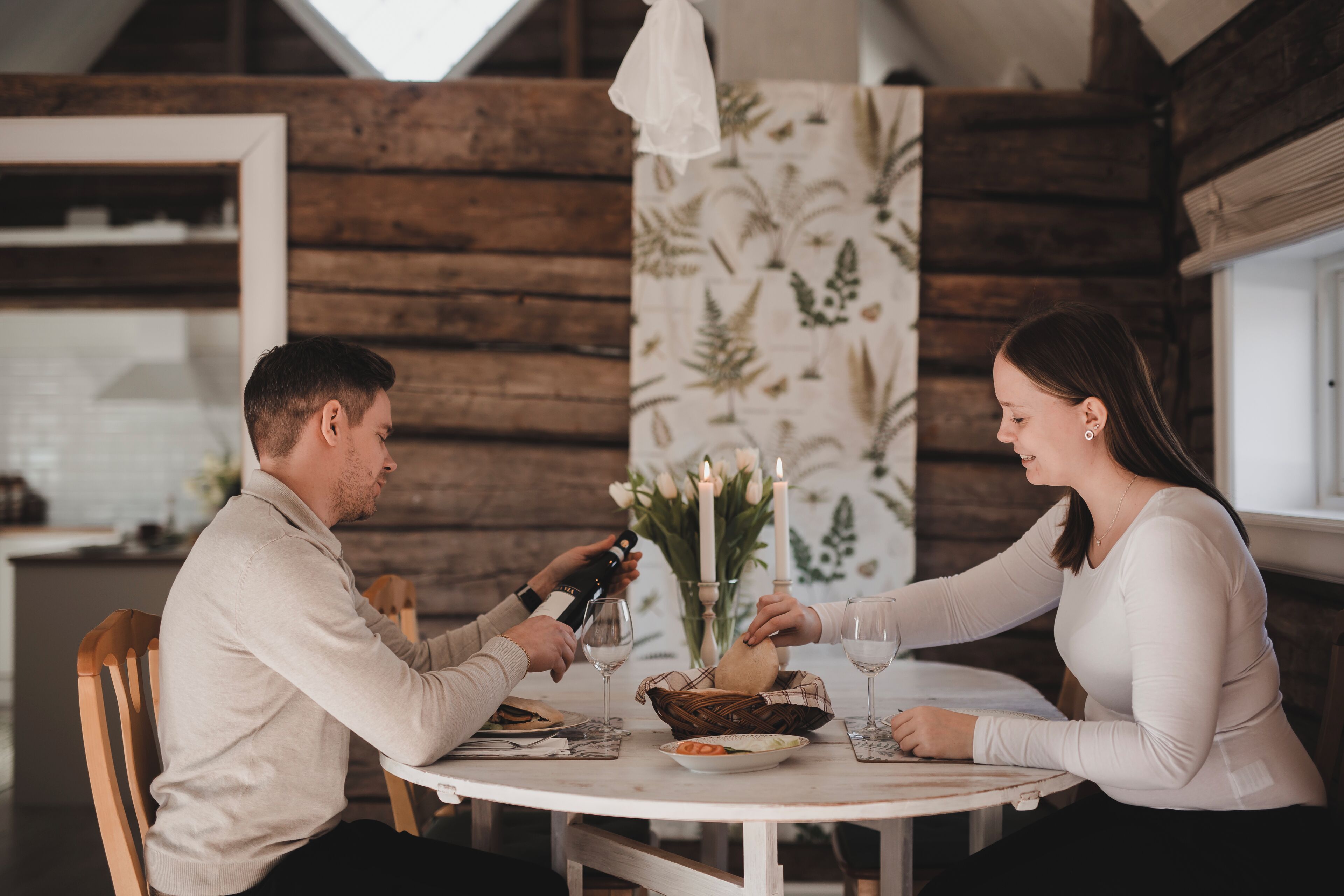 Una pareja sentada a una mesa redonda con mantel blanco, rodeada de paredes de madera y papel tapiz floral.