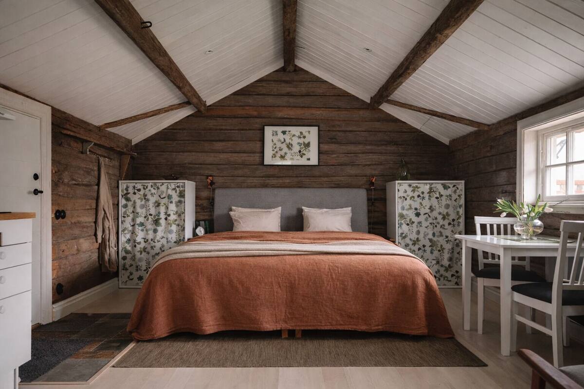 A cozy bedroom with a wooden ceiling, a bed with a brown blanket, floral wallpaper, and a small table with chairs.