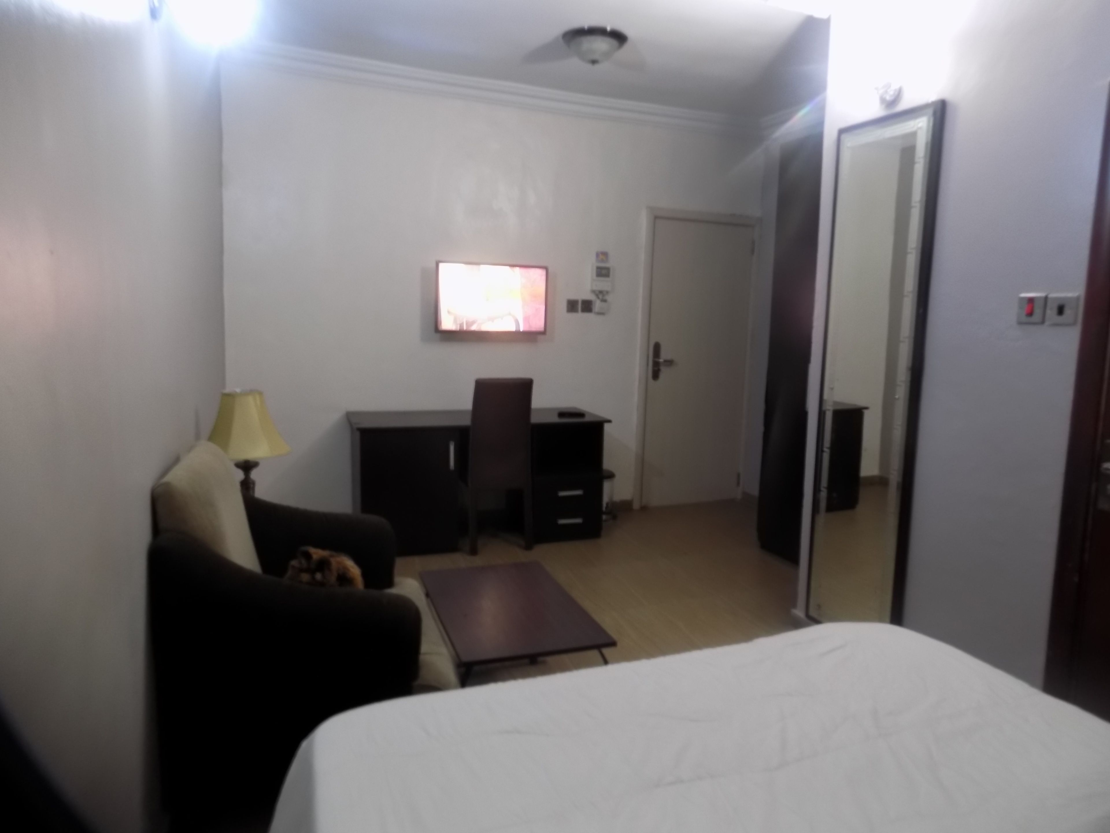 A hotel room with a bed, a desk, a chair, a lamp, a small table, and a door.
