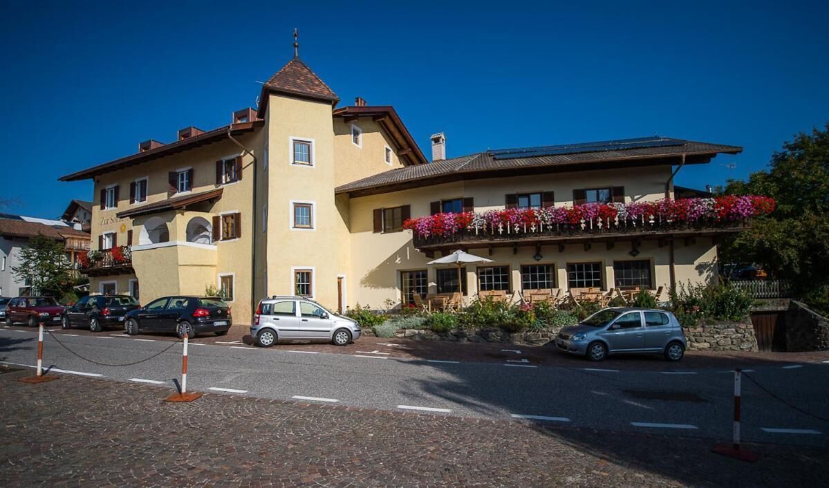 Ein mehrstöckiges Gebäude mit einem an Blumen bepflanzten Balkon, blauem Himmel und parkenden Autos davor.