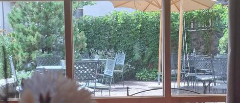 A view through a window with a table and chairs outside, a large umbrella, and greenery.