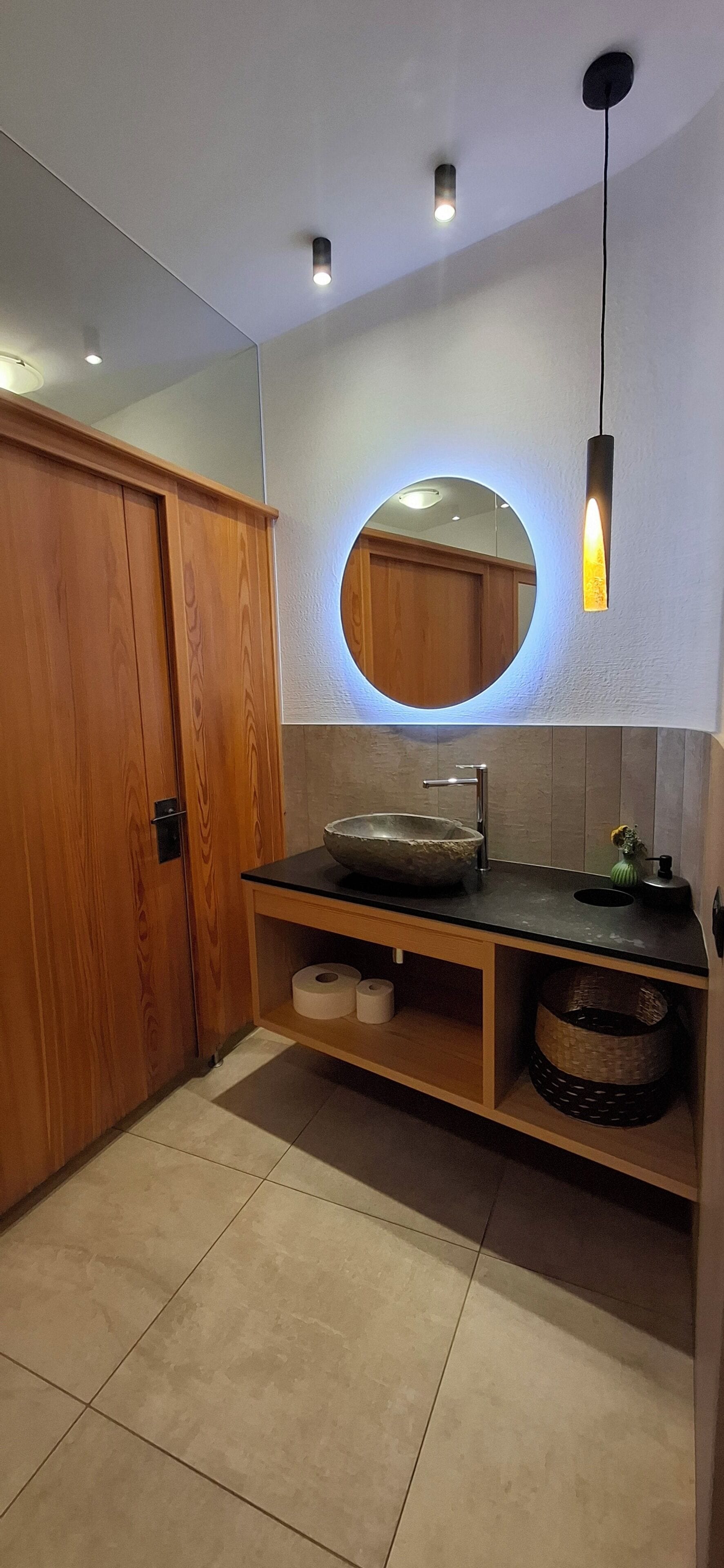 Un baño con un armario de madera, un espejo redondo con luz, un lavabo con pila de piedra y un estante con toallas y cestas.