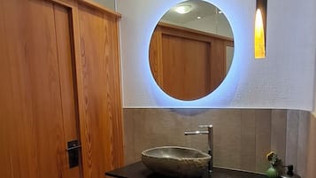 Un baño con un armario de madera, un espejo redondo con luz, un lavabo con pila de piedra y un estante con toallas y cestas.