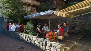 Un groupe de personnes est assis à une table en plein air dans un restaurant dont les murs sont en pierre et qui possède un balcon en bois.