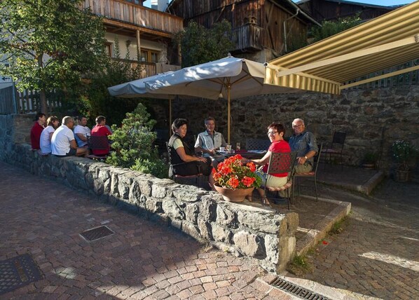 Un groupe de personnes est assis à une table en plein air dans un restaurant dont les murs sont en pierre et qui possède un balcon en bois.