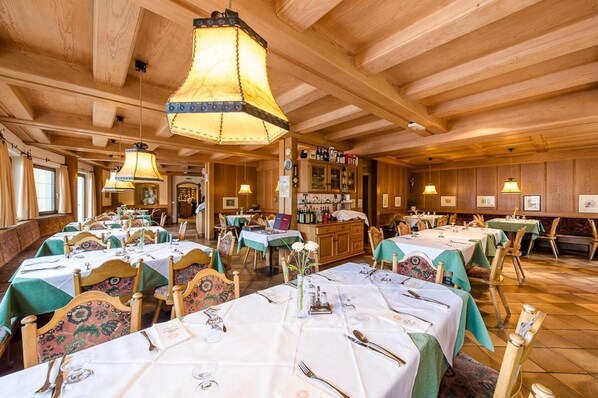 Ein Restaurant mit Holzvertäfelungen, mehreren Tischen mit weißen Tischdecken und grünen Tischläufern, sowie hängenden Lampen.