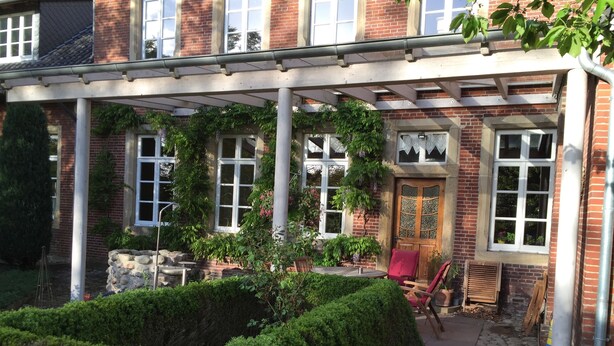 A covered outdoor area with a stone staircase leading to a wooden door, surrounded by greenery and potted plants. - Hotel Cramer (Werlte)