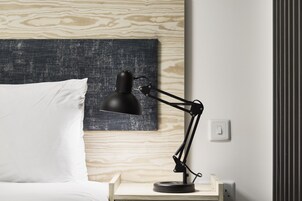 Una cama con almohada blanca y cabecera de madera, lámpara de escritorio negra y un enchufe en la pared.