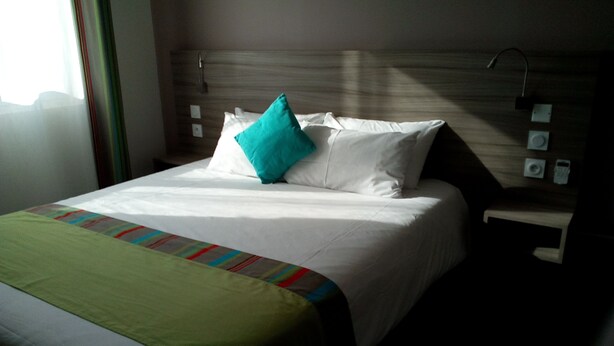Une chambre d’hôtel comprenant un lit, une table de chevet, un panneau de commande fixé au mur et une fenêtre avec des rideaux.