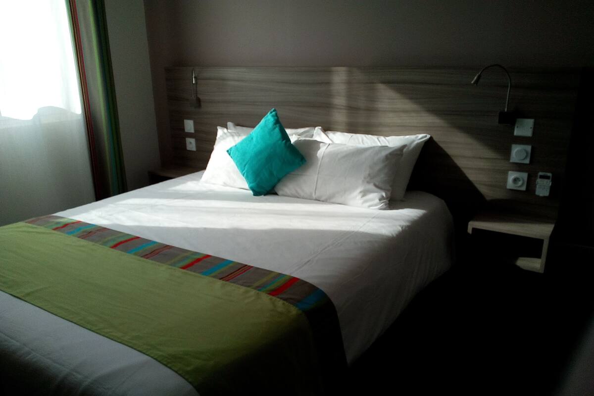 A hotel room with a bed, a nightstand, a wall-mounted control panel, and a window with curtains.
