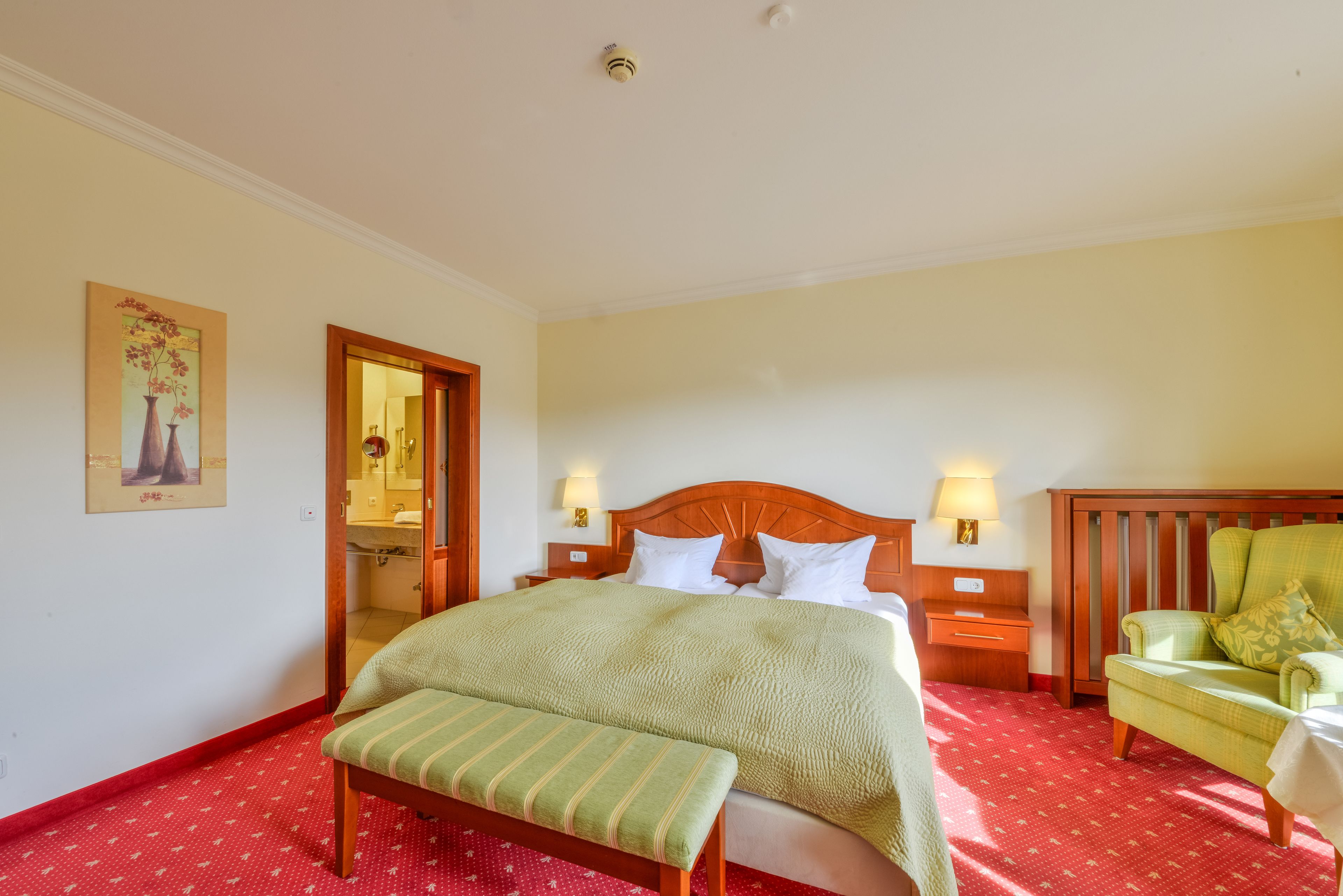 Une chambre d’hôtel comprenant un lit, une banquette, une chaise, une table de chevet et une lampe fixée au mur.