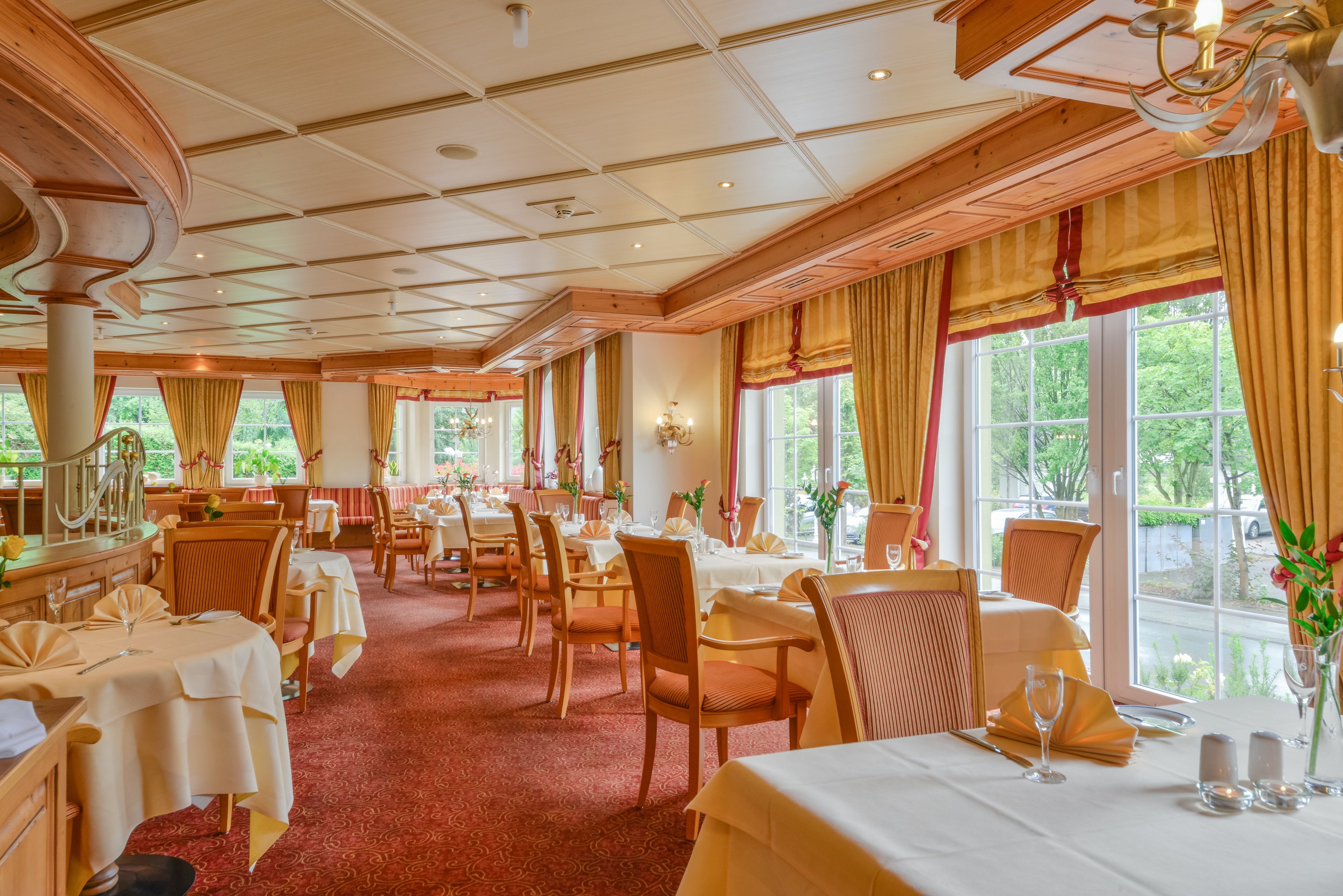 A spacious dining area with multiple tables and chairs, large windows with curtains, and a wooden ceiling.
