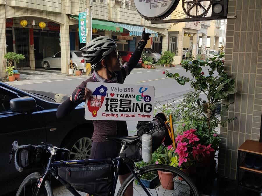 A cyclist holding a sign for Velo B&B Cycling Around Taiwan, standing next to a parked bicycle.