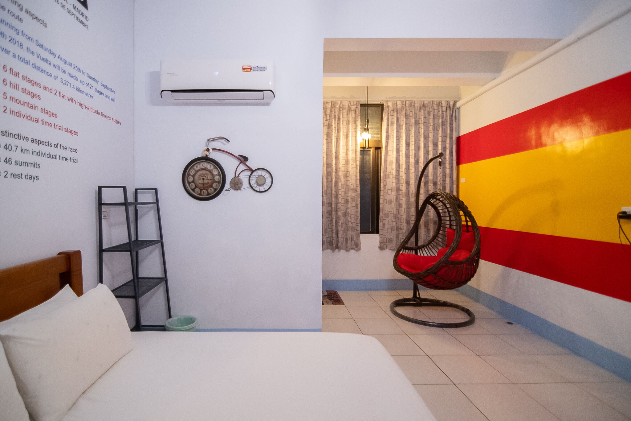 Une pièce comprenant un lit, un climatiseur fixé au mur, un vélo, une chaise en osier et un mur aux couleurs vives.