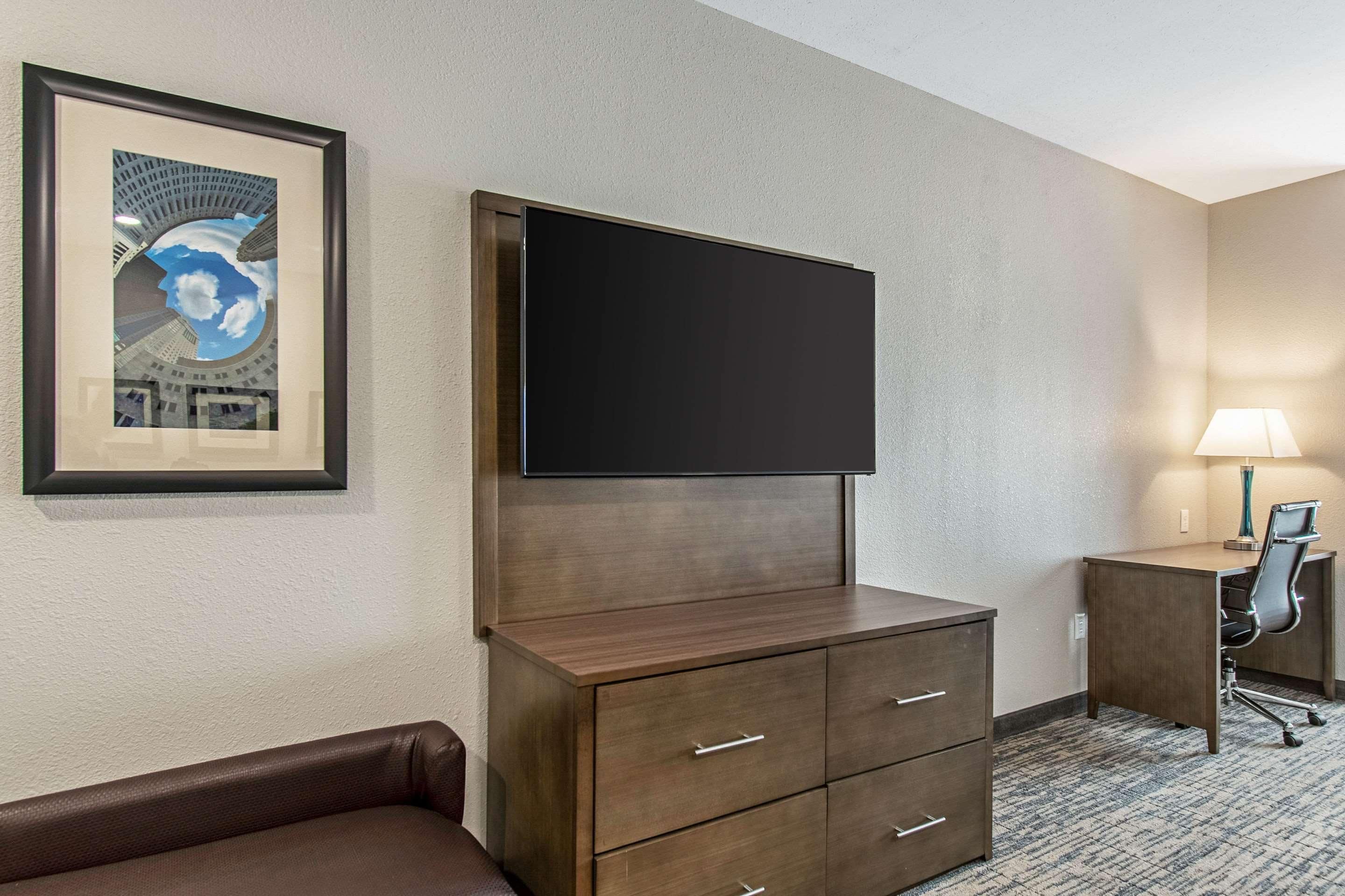 Habitación de hotel con televisor de pantalla plana montado en un mueble de madera, un cuadro en la pared, un escritorio con lámpara y un sofá de cuero marrón.