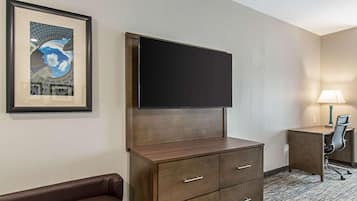 Habitación de hotel con televisor de pantalla plana montado en un mueble de madera, un cuadro en la pared, un escritorio con lámpara y un sofá de cuero marrón.
