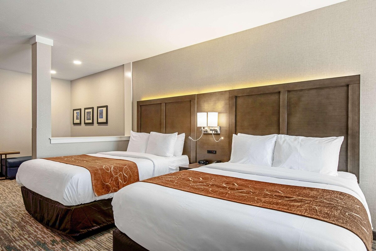 Ein Hotelzimmer mit zwei Betten, einem mit Beleuchtung versehenen Kopfteil, einem kleinen Schreibtisch und einer Wanduhr.