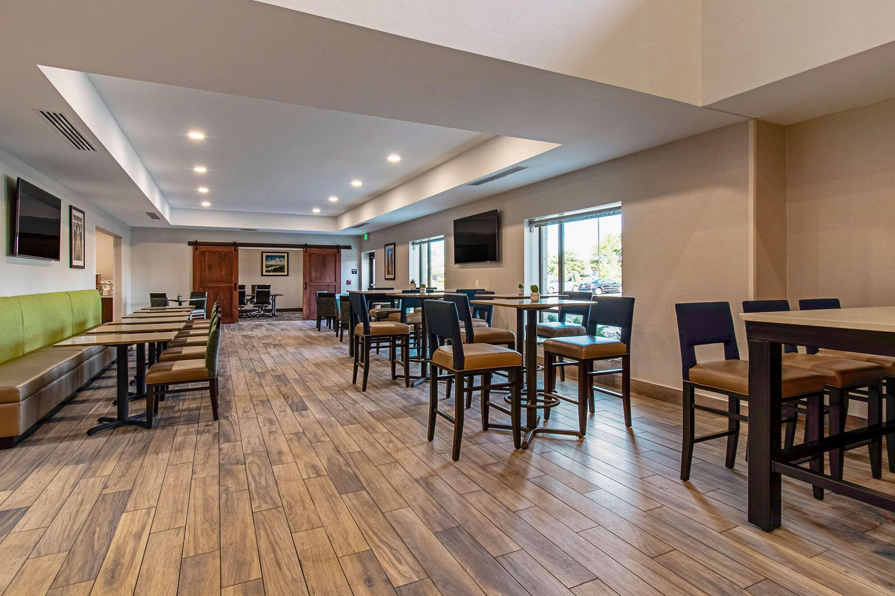 A spacious dining area with wooden flooring, multiple tables and chairs, and large windows providing natural light.