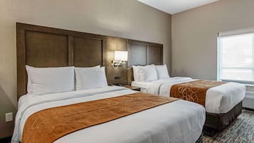 Quarto de hotel com duas camas, uma mesa de cabeceira, uma luminária na parede e uma janela grande.