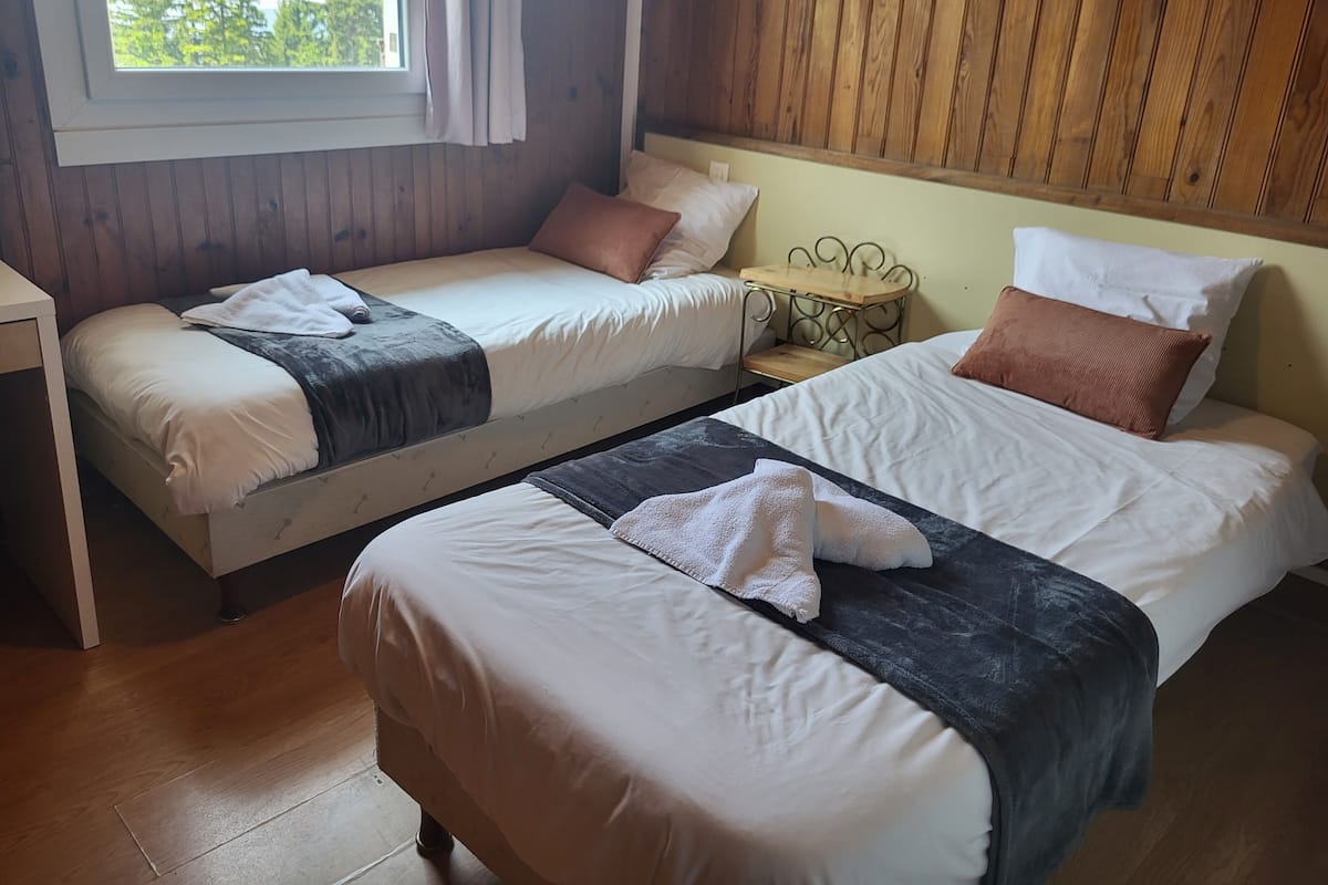 A room with two beds, wooden walls, a window with curtains, and a view of trees.