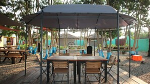 A covered outdoor seating area with wooden tables and chairs, surrounded by trees and a playground in the background. - Tom's Garden (Senya Beraku)