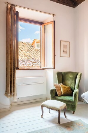 A room with a window, a radiator, a green armchair with a cushion, a small wooden bench, and a patterned rug.
