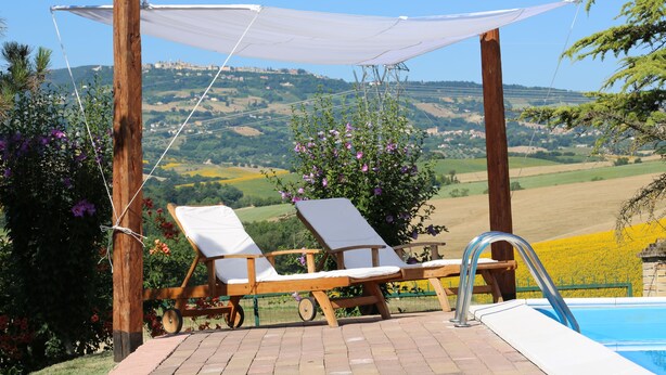 Uma área de piscina com duas espreguiçadeiras e um toldo, com vista para uma paisagem deslumbrante com colinas e um castelo ao longe.