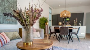 Un salon moderne avec une table basse ronde en bois, un vase en verre contenant des fleurs séchées, un canapé agrémenté de coussins à motifs et un coin repas avec une table et des chaises en bois.