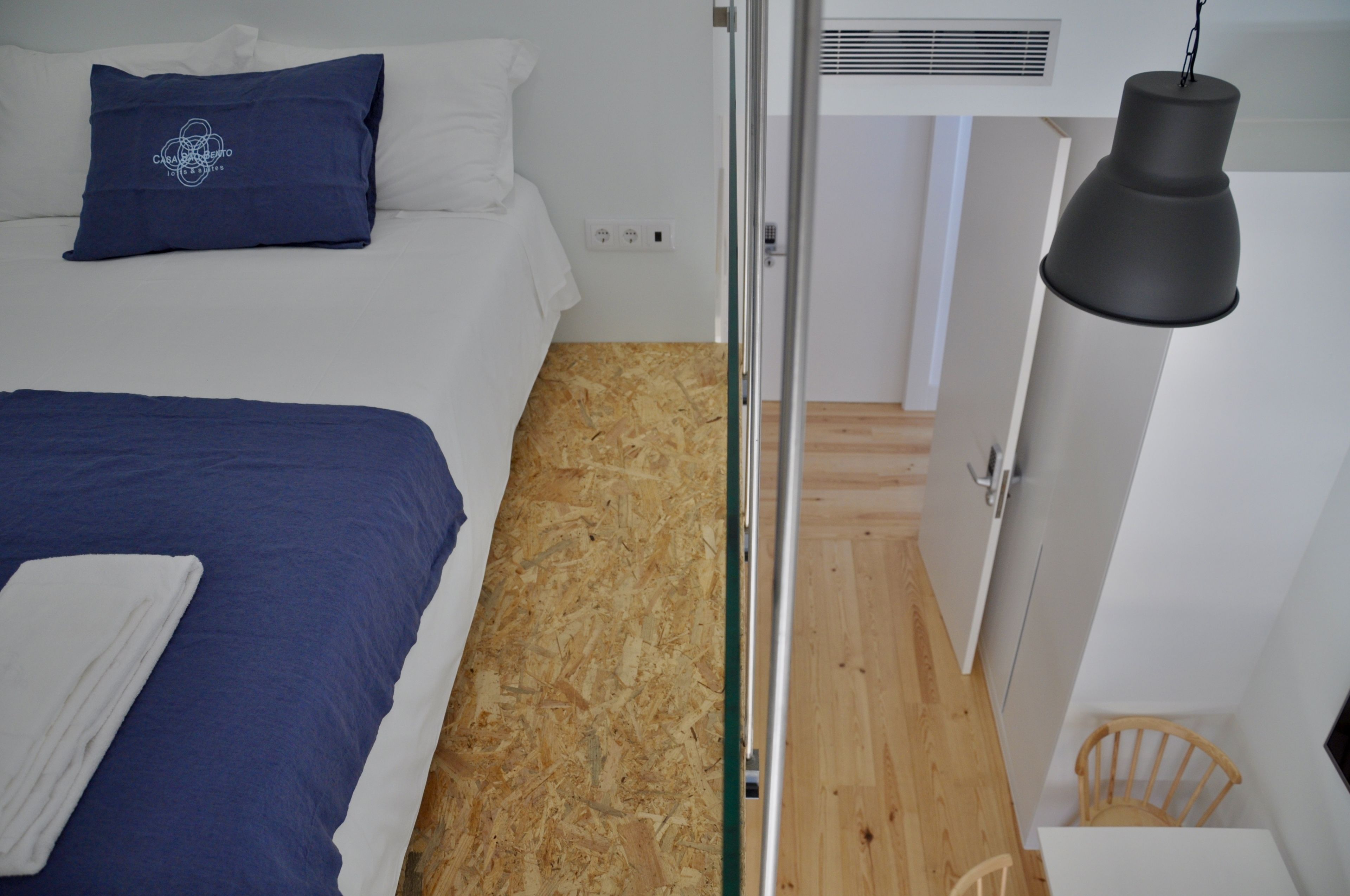 A bedroom with a bed, a blue pillow, a white pillow, a wooden floor, a white wall, a white door, a white ceiling, a black hanging light, and a white chair.