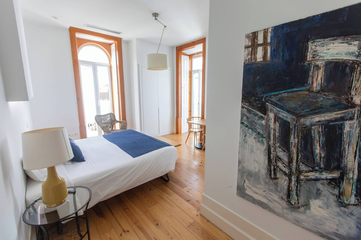 A bedroom with a bed, a lamp, a chair, a table, and a painting.