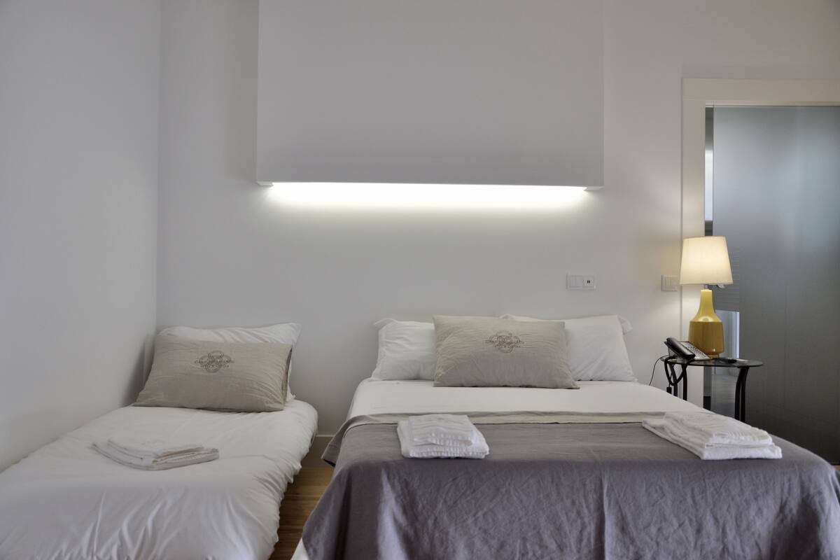 A hotel room with two single beds, a bedside table with a lamp, and a door in the background.