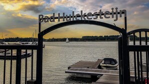 A view through a metal gate with the text "Bad Hülkesteejn" above it, leading to a body of water with boats and a sailboat in the distance.