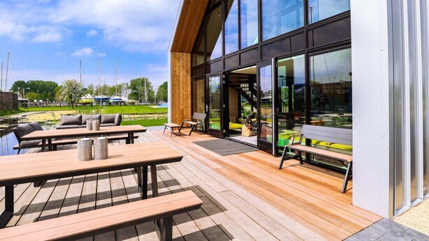 A modern building with large glass windows, a wooden deck with outdoor seating, and a view of boats on a water body. - EuroParcs Bad Hulckesteijn (Nijkerk)