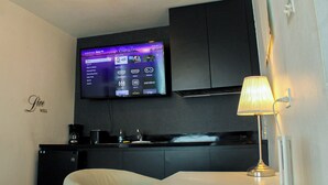 A hotel room with a flat-screen TV mounted on the wall displaying a Roku TV interface, a bed with a white headboard, a bedside table with a lamp, and a dark-colored wardrobe.