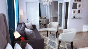 Ein modernes Wohnzimmer mit einem dunklen Sofa, weißen Sessel und einem Kronleuchter, sowie einem gemusterten Teppich.