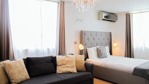 A hotel room with a grey sofa, a bed with a grey headboard, and a chandelier. - Chandelier 202 (Ponce)