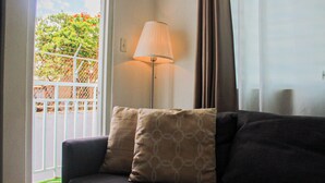 A living room with a dark-colored sofa, two patterned cushions, a floor lamp, and a view of a white gate and greenery outside. - Chandelier 202 (Ponce)