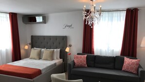 Une chambre d’hôtel moderne, équipée d’un lit, d’un canapé, de deux fauteuils, d’un lustre et d’un téléviseur fixé au mur.