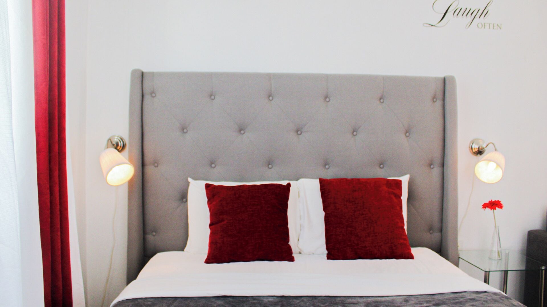 A neatly made bed with a tufted headboard, two red pillows, and a white bedspread. There are two bedside lamps on either side of the bed.
