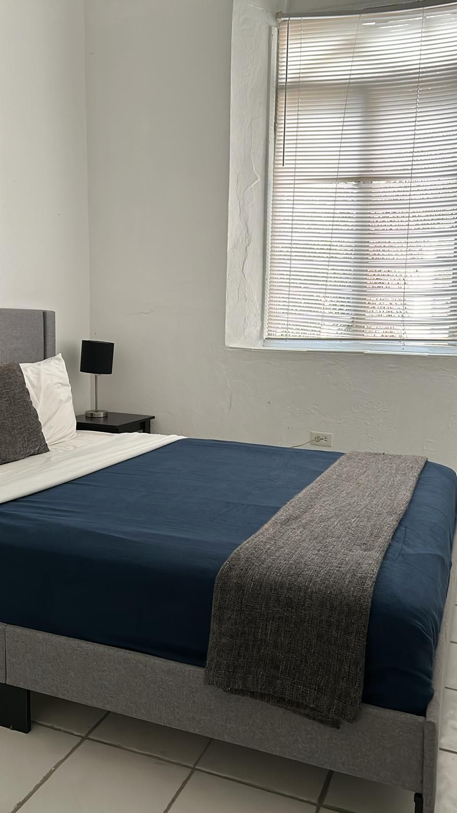 A bedroom with a bed, a nightstand, a lamp, and a window with blinds.