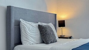 Una cama bien hecha con almohadas blancas y grises, un cabecero gris y una mesita de noche con una lámpara.