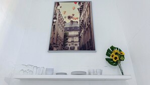Una obra de arte enmarco que muestra un edificio con balcones y globos aerostáticos, un estante blanco con cristalería y una planta en maceta con flores amarillas.