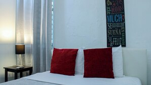 A bedroom with a bed, two red pillows, a white headboard, a nightstand with a lamp, and a wall with a decorative sign.